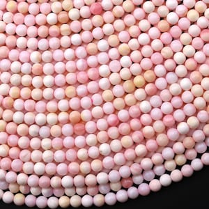 Puede incluir: Una colección de pequeñas cuentas esféricas en varios tonos de rosa suave, crema y melocotón pálido. Cada cuenta presenta un acabado liso y mate con sutiles variaciones naturales en el color y la textura, dispuestas en un patrón denso y uniforme.