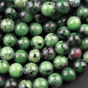 May include: A strand of green, black and pink gemstone beads. The beads are round and have a speckled appearance.