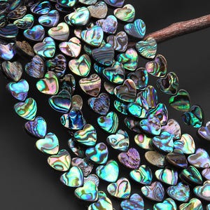 May include: A collection of iridescent heart-shaped beads in shades of blue, green, purple, and brown. The beads are strung together, creating a shimmering effect. The beads are resting on a dark surface.