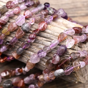May include: A strand of multi-coloured gemstone beads. The beads are a mix of purple, pink, brown, and clear, and they are all tumbled into a smooth, rounded shape. The beads are strung on a clear elastic cord.