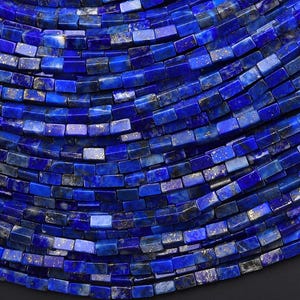 May include: A collection of small, rectangular lapis lazuli beads arranged in multiple curved rows. Each bead features a deep royal blue color with natural inclusions of gold-colored pyrite flecks, creating a textured and vibrant appearance.