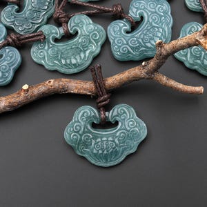 May include: A collection of translucent, blue-green jade-like pendants shaped like traditional locks with intricate carved swirling patterns. Each pendant is attached to a dark brown cord and hangs from a textured, gnarled wooden branch.