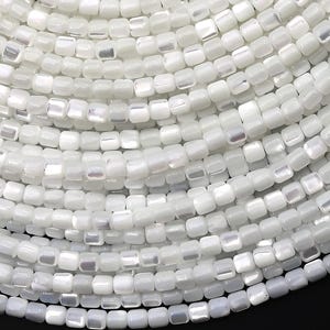 May include: Close-up of a collection of white, square-shaped beads. The beads are strung together, creating a layered, circular pattern. The beads have a pearlescent sheen, reflecting light. The background is black.