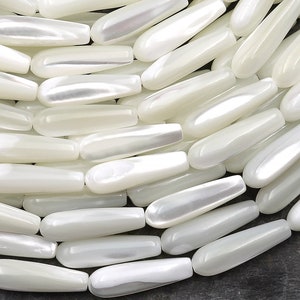 Puede incluir: Primer plano de un montón de cuentas blancas nacaradas en forma de lágrima. Las cuentas son lisas y brillantes, con un ligero brillo iridiscente.