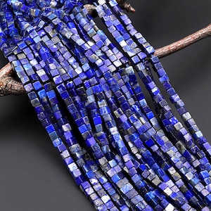May include: Strands of square-cut, deep blue lapis lazuli beads. The beads are a mix of solid and mottled blue hues, creating a textured appearance. The beads are strung together, and are displayed against a dark background.