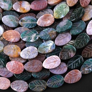 May include: A close-up of oval gemstone beads, each featuring a carved leaf design. The beads display a range of colours, such as green, red, and brown. These beads are ideal for crafting jewellery. The measurements are in centimetres.