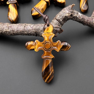 May include: A collection of tiger's eye gemstone cross pendants. Each cross features a brown cord for wearing. The crosses are a mix of golden brown and dark brown, with a polished finish. The crosses are displayed on a dark gray background.