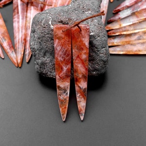 May include: Two long, pointed, red and clear quartz crystal pendants with a rough, natural finish. The pendants are sitting on a dark gray rock.