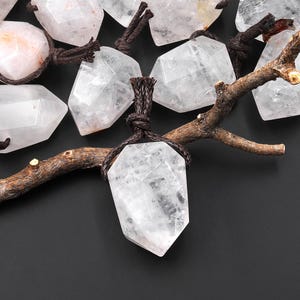 May include: A collection of faceted clear quartz pendants, each suspended by a dark brown braided cord. The stones are irregularly shaped with sharp angles and internal inclusions, arranged around a textured brown wooden branch.