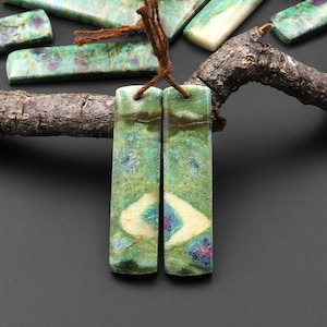 Könnte beinhalten: Ein Paar grüne, weiße und rosafarbene Edelsteinohrringe mit einem einzigartigen Muster. Die Ohrringe sind mit braunem Garn zusammengebunden.