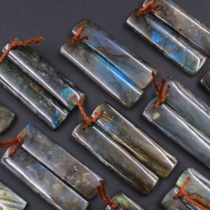 May include: A set of 10 pairs of labradorite gemstone pendants. Each pendant is a double-sided, rectangular shape with a dark gray, blue, and green iridescent color. The pendants are tied together with brown string.