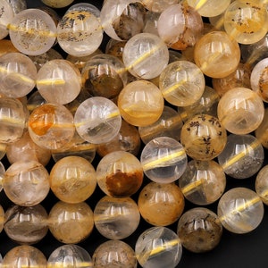 Puede incluir: Una hebra de cuentas redondas de cuarzo rutilado dorado translúcido. Las cuentas tienen una textura natural y rugosa y miden aproximadamente 10 mm de diámetro.