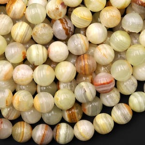Può includere: Primo piano di una collezione di perline rotonde e lucide. Le perline sono principalmente di colore bianco crema, con vortici e motivi gialli, arancioni e marroni. Le perline sono probabilmente realizzate in pietra naturale o minerale.