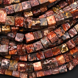 Könnte beinhalten: Nahaufnahme einer Sammlung quadratischer Perlen in Braun-, Rot- und Orangetönen. Die Perlen haben ein marmoriertes Aussehen mit Farb- und Musterunterschieden. Die Perlen sind aufgefädelt, was darauf hindeutet, dass sie für die Schmuckherstellung bestimmt sind.