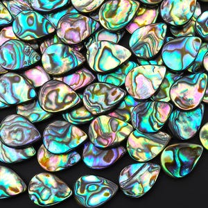 May include: A collection of iridescent abalone shell teardrop-shaped beads. The shells display a range of colors, including blues, greens, purples, and golds, with swirling patterns. The beads are arranged closely together, filling the frame.
