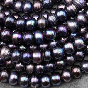 May include: A close-up view of a collection of dark, lustrous, oval-shaped pearls. The pearls have a deep, iridescent sheen, with hints of purple, blue, and green. The pearls are arranged closely together, suggesting they are strung or ready for jewelry making.