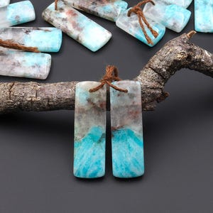 Puede incluir: Colgantes rectangulares de piedras preciosas en tonos azules, grises y marrones, con un aspecto veteado natural. Cada colgante está ensartado con un cordón marrón. Los colgantes se exhiben sobre una superficie oscura y una rama de madera.