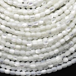 May include: Close-up of a collection of white, rectangular beads. The beads are strung together, creating a layered, textured appearance. The beads have a pearlescent sheen, reflecting light and adding a subtle shimmer to the overall aesthetic.