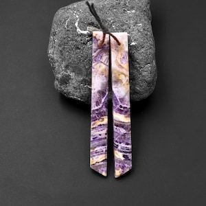 May include: Two long, thin, purple and white striped gemstone pendants with a pointed end. The pendants are on a dark gray surface.