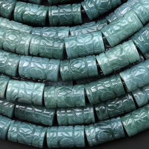 Puede incluir: Primer plano de un collar de cuentas de jade talladas. Las cuentas son cilíndricas, con intrincados diseños florales y geométricos. El jade tiene un color verde-azulado translúcido, y las cuentas están ensartadas en un hilo oscuro, creando un collar.
