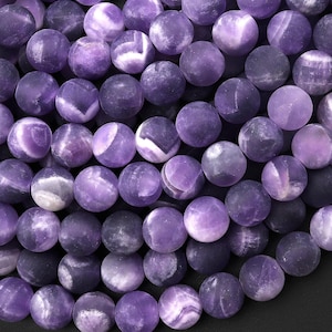 May include: A close-up of a strand of purple amethyst beads. The beads are round and have a smooth, polished finish. The amethyst is a light purple color with white streaks throughout.