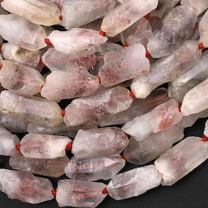 May include: A collection of raw, translucent quartz crystal beads strung together on a thin red cord. Each bead features an irregular, elongated shape with natural inclusions, displaying shades of white, pale pink, and grey throughout the stones.