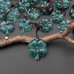 May include: A collection of translucent, teal-colored four-leaf clover pendants with brown cords. The pendants are arranged on a dark surface with a brown branch, creating a natural and elegant display. The clover pendants are a symbol of good luck.