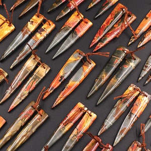 May include: A set of 12 pairs of gemstone earrings. Each pair features two long, pointed, teardrop-shaped stones with a brown, orange, and gray marbled pattern. The stones are attached to a brown cord with a knot at the top.
