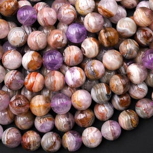 May include: A collection of round, polished gemstone beads featuring a marbled pattern of soft lavender, deep purple, and earthy brown tones. Each bead displays unique swirling bands and translucent areas, creating a smooth, glossy surface.