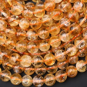 May include: A close-up shot of a collection of golden-yellow citrine gemstone beads. The round, translucent beads are strung together with orange thread. The beads have a warm, amber hue, and are suitable for jewelry making.