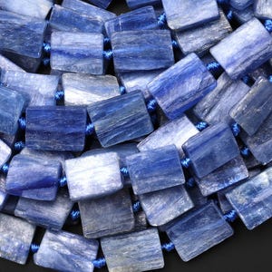 Puede incluir: Primer plano de un collar hecho de cuentas rectangulares de kianita azul. Las cuentas var&iacute;an en tonos de azul, de claro a &iacute;ndigo profundo, y est&aacute;n ensartadas en un cord&oacute;n azul. La piedra natural tiene una apariencia texturizada.