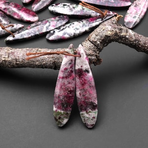 May include: Two long, teardrop-shaped, pink and green gemstone pendants with a white speckled pattern. The pendants are hanging from a brown branch.