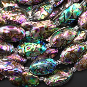 Könnte beinhalten: Nahaufnahme einer Sammlung schillernder Abalone-Muschelkernperlen. Die oval geformten Perlen zeigen eine Reihe von Farben, darunter Lila, Grün und Gold, mit einem glänzenden Finish. Jede Perle ist mit einem kleinen, transparenten Abstandshalter aufgefädelt.