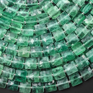 May include: Close-up of a necklace made of faceted, square-cut green gemstones. The gems are strung together with small silver-colored beads. The necklace has a layered appearance, with multiple strands visible.