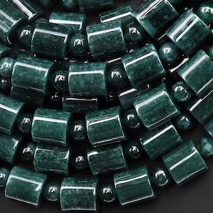 May include: Close-up of a dark green necklace featuring cylindrical and spherical beads. The beads have a polished, glossy finish, reflecting light. The necklace is likely made of jade or a similar gemstone, with a natural, organic aesthetic.