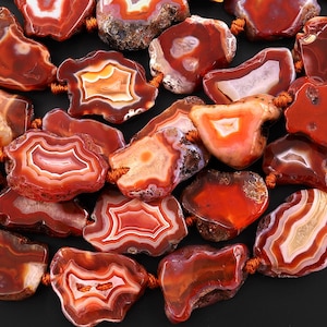 Puede incluir: Una colección de cuentas de ágata pulidas en tonos rojos, naranjas y blancos. Las cuentas, de forma irregular, están ensartadas con un cordón marrón, mostrando patrones y texturas naturales. Ideal para la creación de joyas.