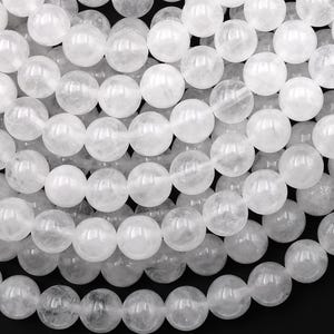May include: Close-up of a collection of translucent, round, white gemstone beads. The beads are strung together, creating a textured surface. The beads are uniform in size and shape, with a glossy finish. The background is black.