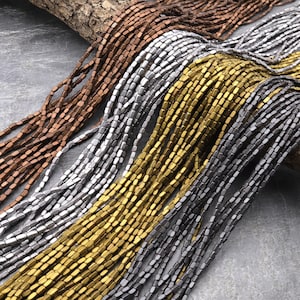 Puede incluir: Una colección de cuatro hebras de cuentas rectangulares facetadas en tonos dorados, plateados y cobrizos. Las cuentas están dispuestas en forma de abanico, con las cuentas doradas en la parte inferior y las cuentas cobrizas en la parte superior.