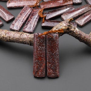 May include: Close-up of rectangular, reddish-brown beads with a textured surface, strung on a brown cord. The beads are arranged on a tree branch, with a dark gray background. The beads have a unique, patterned appearance.