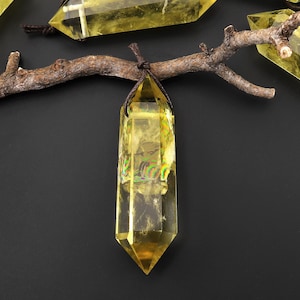 May include: A yellow crystal point pendant with a brown leather cord. The crystal is clear and has a slight iridescent sheen. The pendant is hanging from a branch.