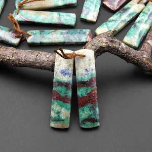 May include: Two long, rectangular, green, white, and brown patterned stones with a brown string attached to each. The stones are on a branch.