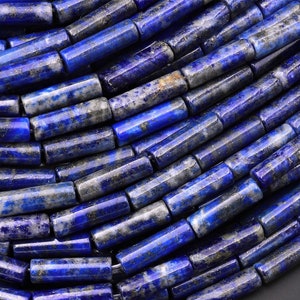 Op de afbeelding: Close-up van een streng lapis lazuli kralen. De kralen zijn cilindrisch, met een diepblauwe kleur en vlekken van goud en grijs. De kralen zijn aan elkaar geregen, waardoor een textuur ontstaat.