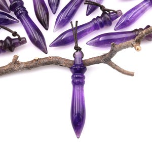 May include: Amethyst crystal pendulums with a pointed tip and a decorative top, each attached to a black cord. The purple gemstones are arranged on a white background with a small tree branch.