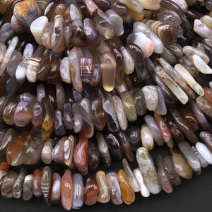 Puede incluir: Primer plano de una hebra de cuentas de ágata. Las cuentas son una mezcla de marrón, blanco y gris con patrones naturales. Las cuentas son lisas y pulidas.