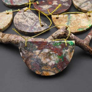 May include: A collection of semi-circular, natural stone ornaments with gold string hangers. The stones display a variety of colors including red, green, brown, and beige. The ornaments are arranged on a dark surface, suggesting a decorative or craft theme.