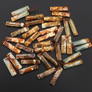 May include: A collection of rectangular gemstone beads in various earth tones, including brown, beige, and green. Each bead has a small hole and is strung with a brown cord. The beads are scattered on a dark surface.