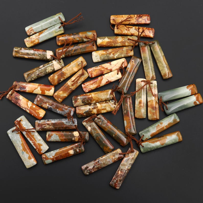May include: A collection of rectangular gemstone beads in various earth tones, including brown, beige, and green. Each bead has a small hole and is strung with a brown cord. The beads are scattered on a dark surface.