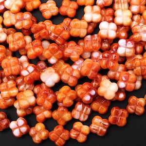 May include: A collection of orange, four-petal flower-shaped beads. The beads have a glossy finish and vary in shades of orange, with some featuring white accents. These beads are likely for jewelry making or crafting.
