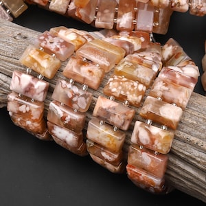 May include: A multi-strand bracelet composed of rectangular, polished flower agate beads. The beads feature translucent, warm-toned hues of peach, tan, and cream, with distinct white, flower-like inclusions throughout the stone surfaces.