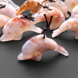 May include: A collection of white agate dolphin pendants with brown cords. The dolphins have a natural, cloudy, and translucent appearance with brown and pink agate inclusions.